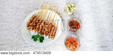 A Plate Of Chicken Satay Served With Lime, A Glass Of Pickles And Sambal Matah In The Room, Their Vi