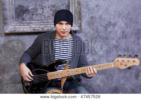 Jeune homme en gilet rayé et veste est assis avec la guitare près de mur éraflé gris