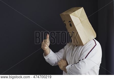 A Man In A White Shirt With A Paper Bag On His Head, With A Smiley Face Drawn, Stands Against The Wa