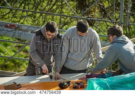 Three Young Carpenters Working On A Roof. A Carpenter With Impact Driver Screws A Screw Into A Woode