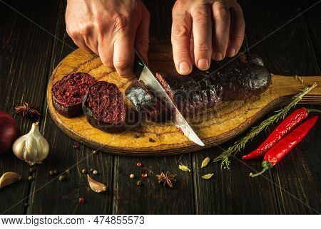 Chef's Hands With A Knife Cut Blood Sausage On A Kitchen Cutting Board. Cooking A National Dish With