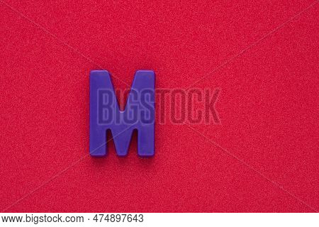 Alphabet Letter M - Violet Colored Plastic Piece On Red 032 C Foamy Background