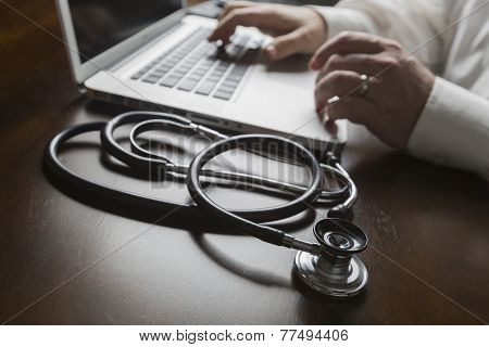 Medical Stethoscope Resting on Desk As Male Hands Type on Computer Keyboard.