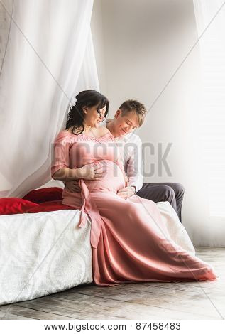 Pregnant Couple Sitting On Bed With Baldachin Next To Window