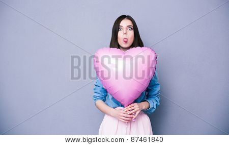 Woman holding heart shaped balloon and showing her tongue over gray background. Looking at camera