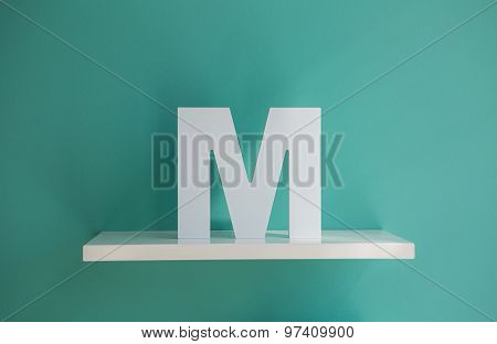 Letter M turquoise color on a white shelf.