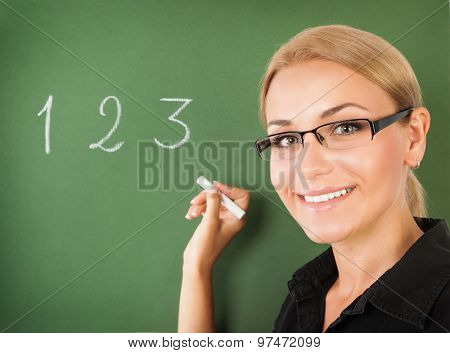 Closeup portrait of young beautiful teacher wearing glasses writing numbers on green chalkboard, start of education season, back to school