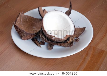 Still life with broken coconut shell ripe white flesh inside and debris on white palte on brown wooden table horizontal close up photo
