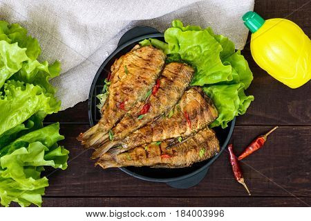 Fried fish carp (sazan) on a cast-iron frying pan with lettuce leaves on a dark wooden background. The top view.