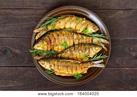 Fried fish carp (sazan) on a ceramic bowl with branches of rosemary on a dark wooden background. The top view.