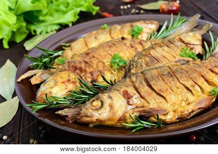 Fried fish carp (sazan) on a ceramic bowl with branches of rosemary on a dark wooden background. Close up