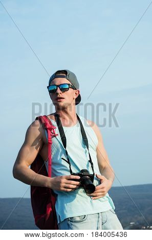 Caucasian man young traveler or photographer with camera in muscular hands triceps biceps in stylish sunglasses and cap red backpack standing on sunny day on blue sky. Leisure and summer vacation