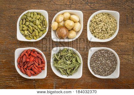 collection of superfoods in small ceramic bowls against rustic wood: pumpkin seeds, macadamia nuts, hemp seed hearts, goji berry, stevia herb and chia seeds