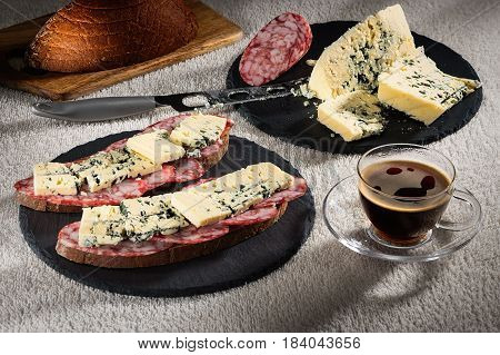 Sandwiches with cheese of Dorblu and raw smoked sausage on black round slate plates, a cheese knife and a cup of hot coffee