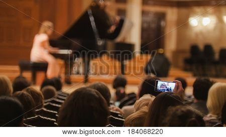 Spectators in concert hall during performing piano girl- people shooting performance on smartphone, telephoto