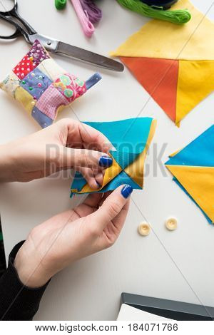 needlework and quilting in the workshop of a tailor on white background - close-up on hands of a tailor holding scraps of fabrics lying on a white table with buttons, pin cushion, scissors