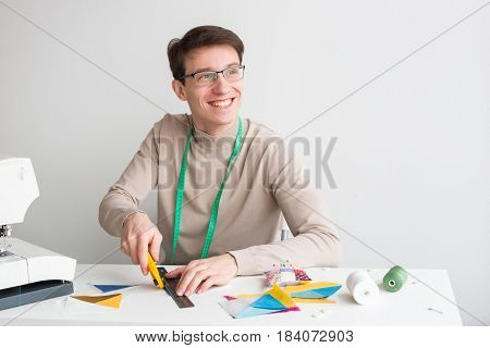 quilting - smiling young male tailor sitting behind a desk and cut out colorful pieces of fabrics for sewing patchwork products, near are spolls of thread, pieces of fabrics and pin cushion