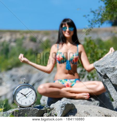 Watches and tanned blurred young woman in bikini sitting in lotus position in a mountainous area . Concept of time to relax. Square frame shallow depth of field.