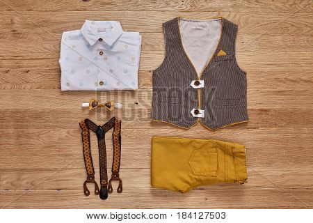 Top view photo of the boy's clothes on the wooden background. White shirt with print, brown vest, bowtie, brown pants and suspenders. Cute clothes for little boys. Concept of the children's fashion. Summer or spring children's clothes.