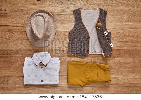 Top view photo of the boy's clothes with accessories on the wooden background. White shirt with print, brown vest, bowtie, brown pants and hat. Cute clothes for little boys. Concept of the children's fashion.