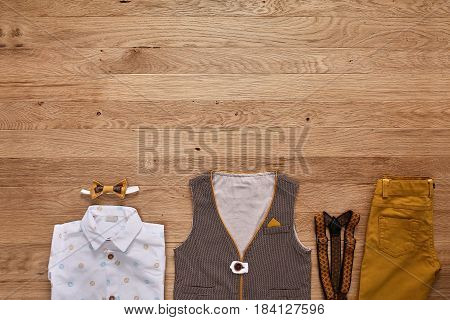 Top view photo of the boy's clothes on the wooden background. White shirt with print, brown vest, bowtie, brown pants and suspenders. Cute clothes for little boys. Concept of the children's fashion. Summer or spring childrens clothes.