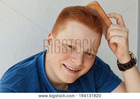 A Smiling Young Attractive Freckled Man With Trendy Hairdo Combing His Red Hair With A Comb Wearing