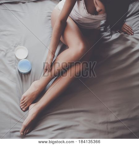 Beautiful woman sitting on bed and applying cream on legs.