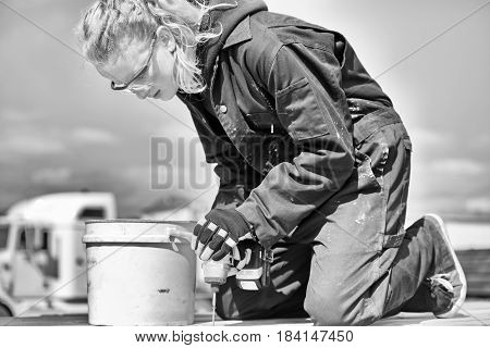 A black and white picture of teenage girl with long hair in a ponytail wearing safety glasses and coveralls drilling a screw into a piece of wood outdoors