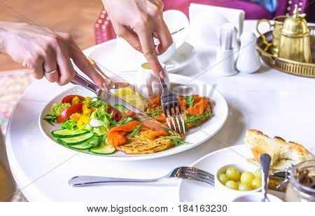 Hands of the girl cuting potato pancakes with sauce. Vegetable fritters. Latkes on white plate, served with salt salmon. Beautiful girl offscreen.