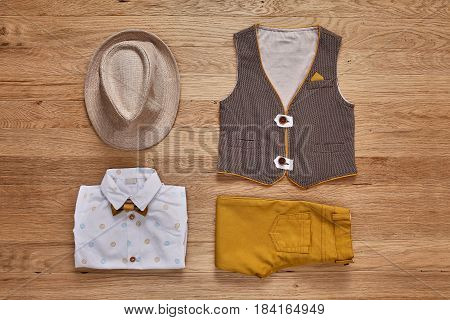 Top view of the kids fashion clothes with accessories on the wooden background. White shirt with print, brown vest, bowtie, brown pants and hat. Elegant clothes for little boys. Concept of the children's fashion.