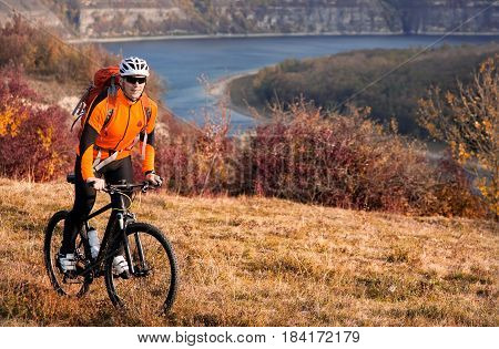 Cyclist in orange sporyive jacket riding the bike on the hill under river against beautiful mountain. Sportsman in the sportwear, helemet, sunglasses and with red backpack. Summer season. Healthy lifestyle.