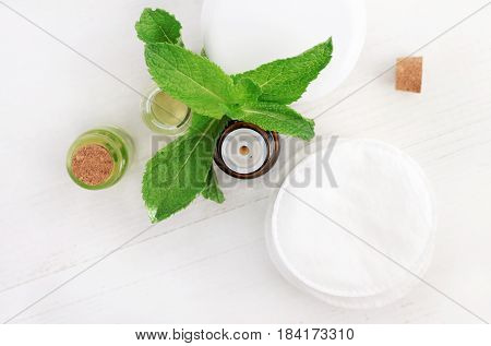 Dropper bottle of essential oil with fresh mint plant leaves for cosmetic use, top view over white wooden table.