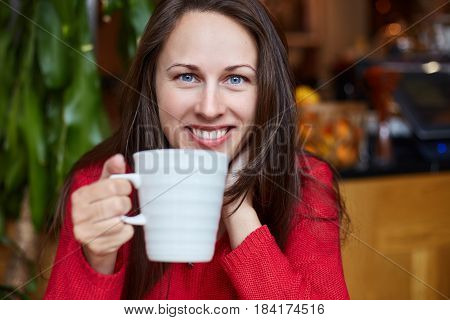 Young natural cheerful Caucasian brunette blue-eyed girl in red sweater is having her breakfast in modern organic coffee shop. Looking and smiling happily at the camera. Youth and beauty concept.