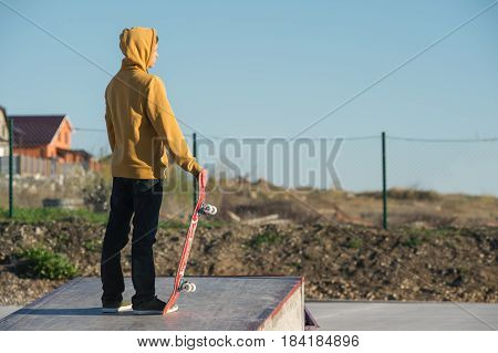 A teenager standing in a yellow hoodie holds a hand skateboard against the backdrop of the outskirts of the city's shantytown and blue sky