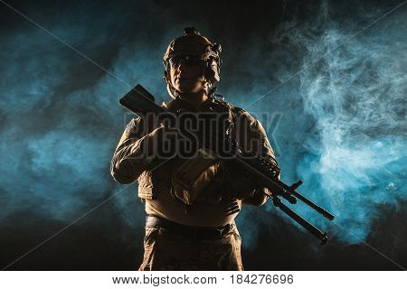 Army soldier in Combat Uniforms with machine gun, plate carrier and combat helmet are on. Studio contour silhouette shot, backlight, dark glowing smoke background