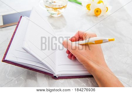 Close up Woman hand writing a 'to do list' in her notebook with background of a cup of fresh herb tea f bouquet of daffodils and her smartphone on the desk. Selective focus.