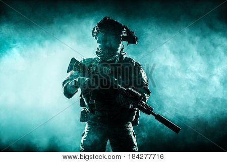 Army soldier in Combat Uniforms with assault rifle, plate carrier and combat helmet are on, Shemagh Kufiya scarf on his neck. Studio contour silhouette shot, backlight, dark glowing smoke background