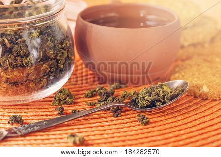 Dry leaves of green tea in a glass jar. Nearby tea leaves in a teaspoon and ceramic tea-things. Composition on a wattled napkin. The cover banks is open several tea leaves are scattered.
