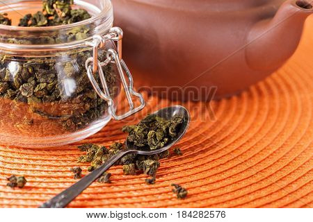 Dry leaves of green tea in a glass jar. Nearby tea leaves in a teaspoon and ceramic tea-things. Composition on a wattled napkin. The cover banks is open several tea leaves are scattered.