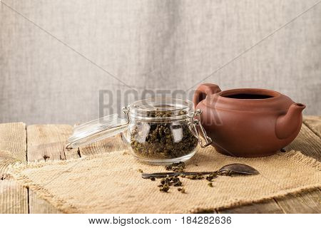 Dry leaves of green tea in a glass jar. Nearby tea leaves in a teaspoon and ceramic tea-things. Composition on a napkin from a sacking. The cover banks is open several tea leaves are scattered. Light background