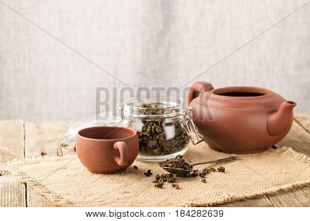Dry leaves of green tea in a glass jar. Nearby tea leaves in a teaspoon and ceramic tea-things. Composition on a napkin from a sacking. The cover banks is open several tea leaves are scattered. Light background