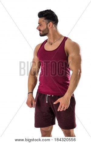 Attractive smiling muscular man in red sleeveless tanktop and shorts, looking to a side at blank space next to him, isolated on white in studio