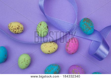 Lilac background with speckled pink green yellow and blue coloured sweet eggs and purple satin ribbon - Easter