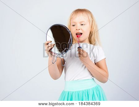 Little happy girl paints lips looking in the mirror. on a white background