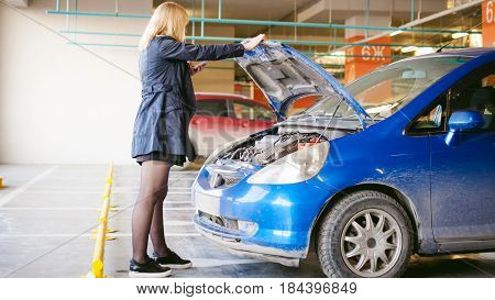 Woman Near Car's Hood. Young Blonde In Covered Parking Of Shopping Center, Stands Near Car With Rais
