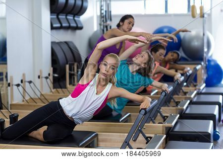 Female trainer assisting group of women with stretching exercise on reformer in gym