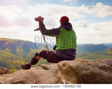 Tired Tourist With Medicine Crutch  And Broken Leg Fixed In Immobilizer Resting On Summit.