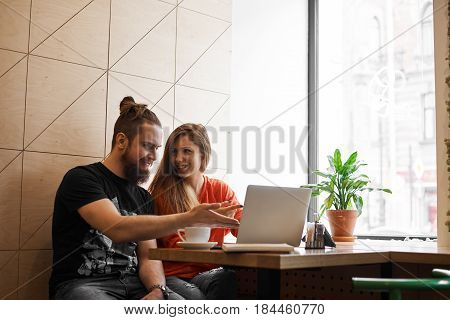 Stylish guys show in laptop and smiling in cafe. Happy man and woman making business plan of trendy online shop. Couple in love typing planning list for weekend with things to do together