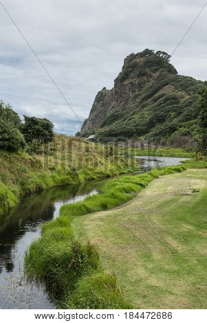 Auckland New Zealand - March 2 2017: Creek between Karekare Waterfall and Karekare beach flows between green lawn and wild area. Forested hill and cloudscape over beach.