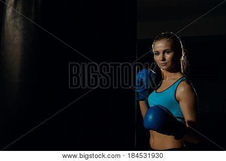 Girl boxer in blue boxing gloves near a punching bag.
